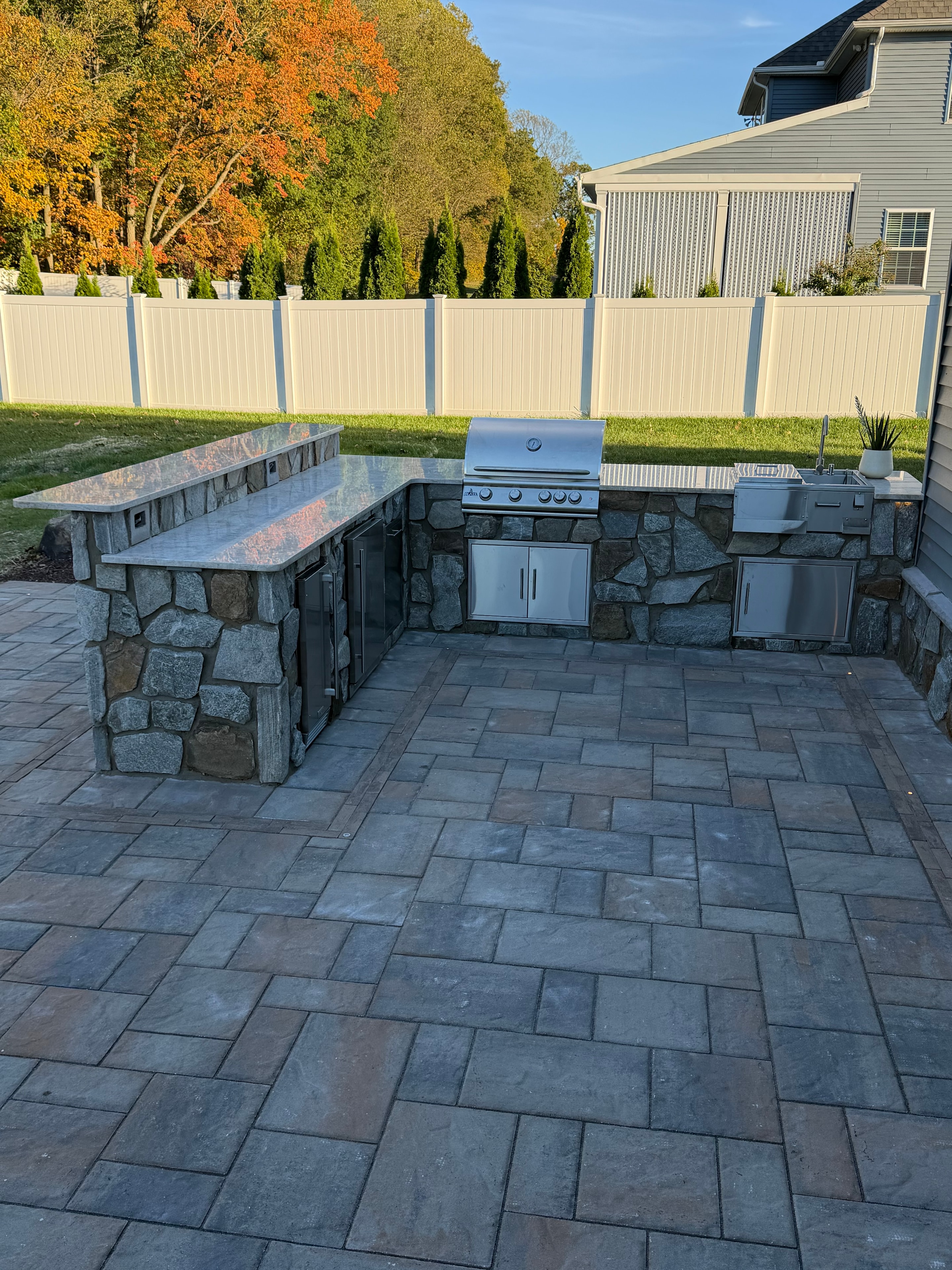 Chef's Dream — daytime close-up of outdoor kitchen with dark stone surround, stainless grill, griddle, and access drawers set into Techo Bloc paver patio with fall foliage backdrop