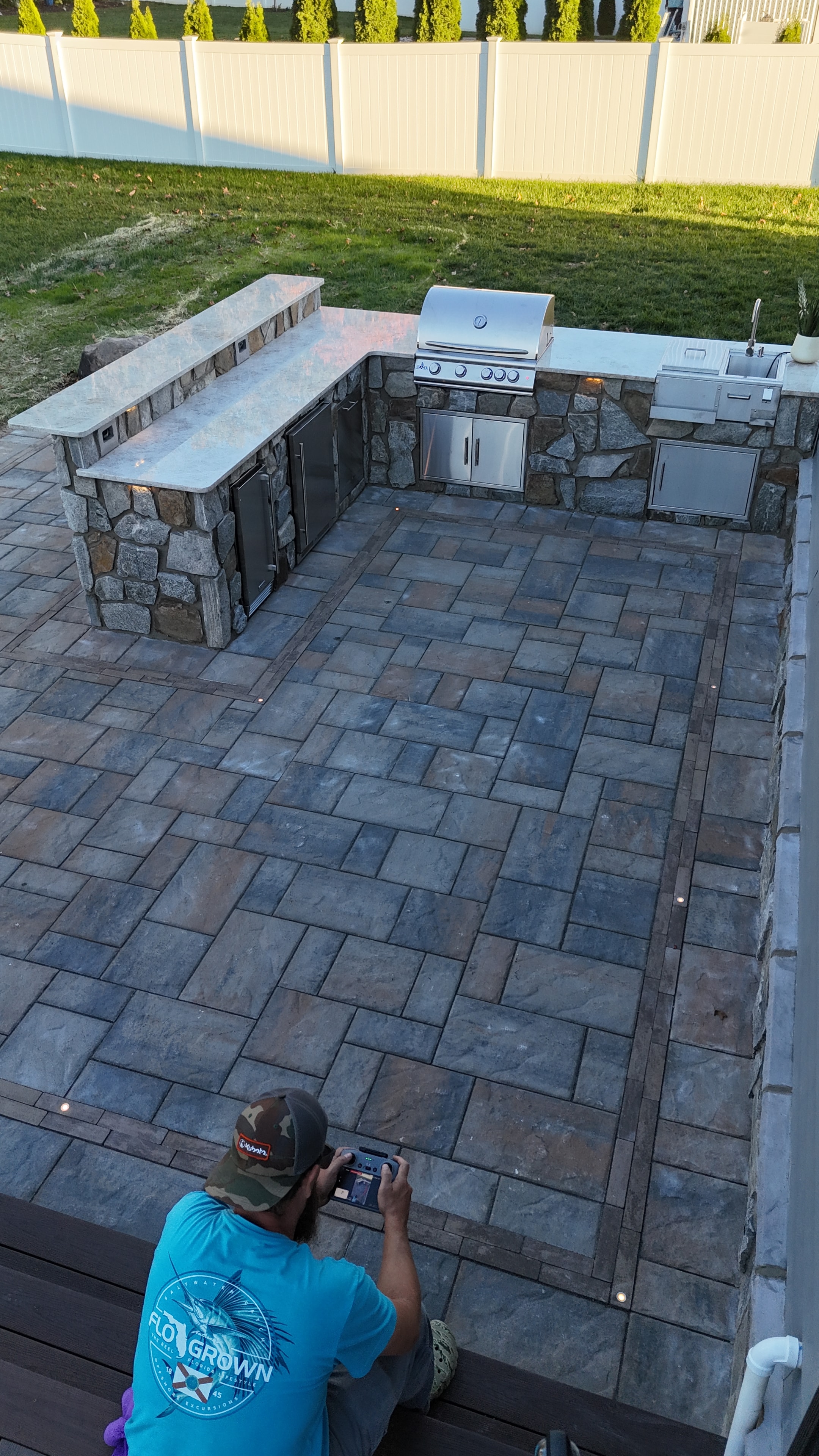 Chef's Dream — aerial angled shot of full paver patio with dining set, composite deck with lounge furniture, outdoor kitchen, boulder border, and green lawn
