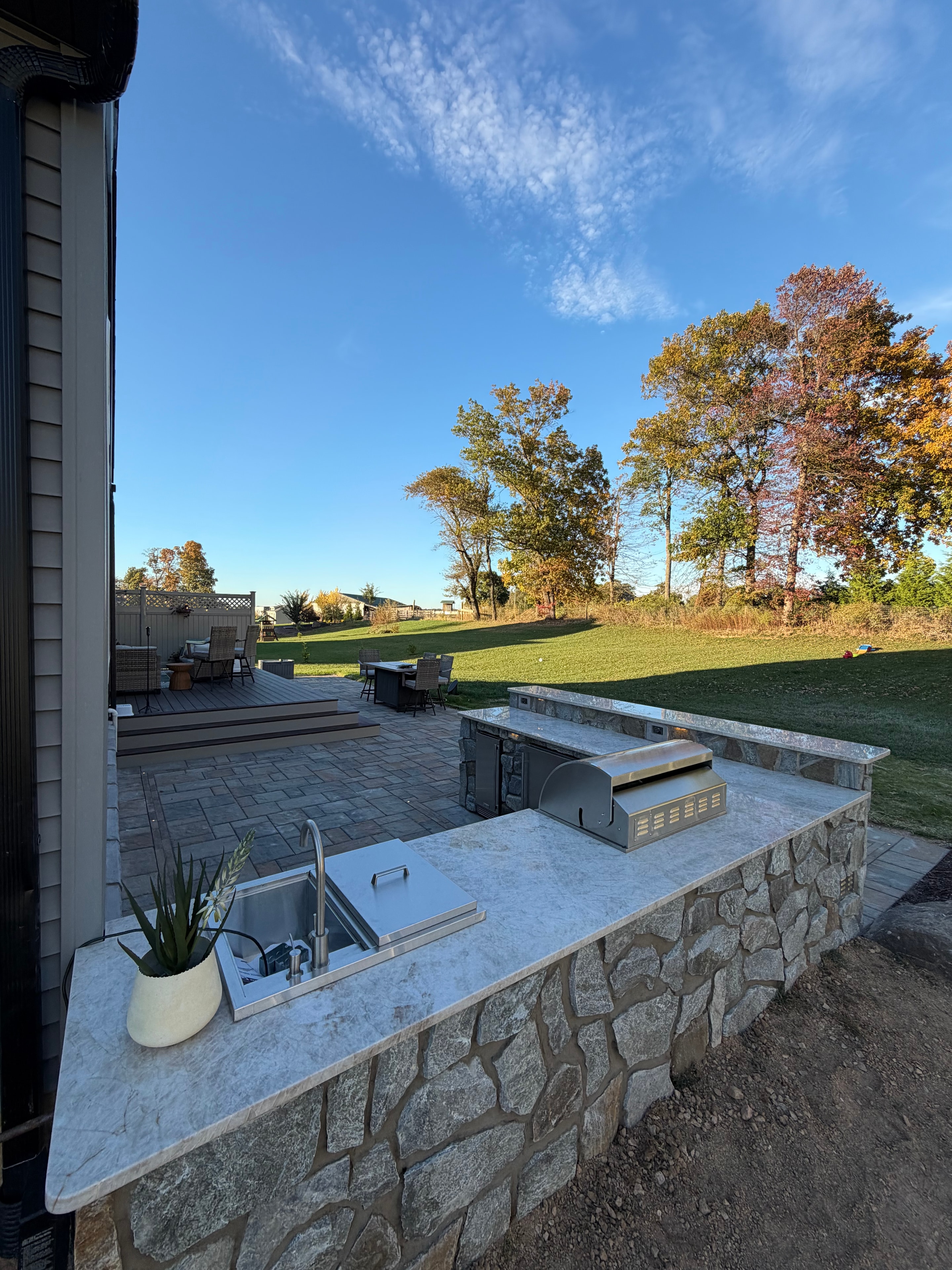 Chef's Dream — daytime ground-level view of granite countertop outdoor kitchen with stainless grill and side burner, stone surround, paver patio, and open green lawn with fall foliage