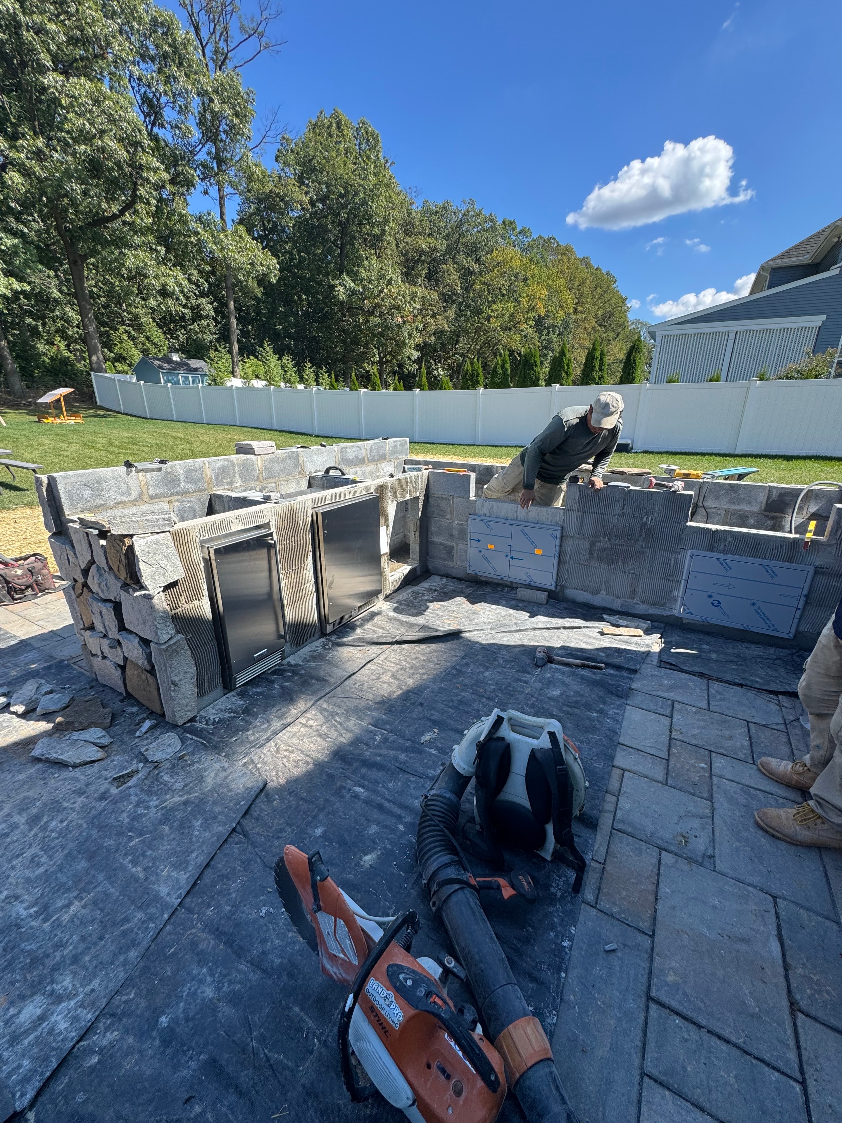 Chef's Dream — crew member directing work on Techo Bloc patio with outdoor kitchen stone surround being built, Stihl cut-off saw and tools on site, green trees in background