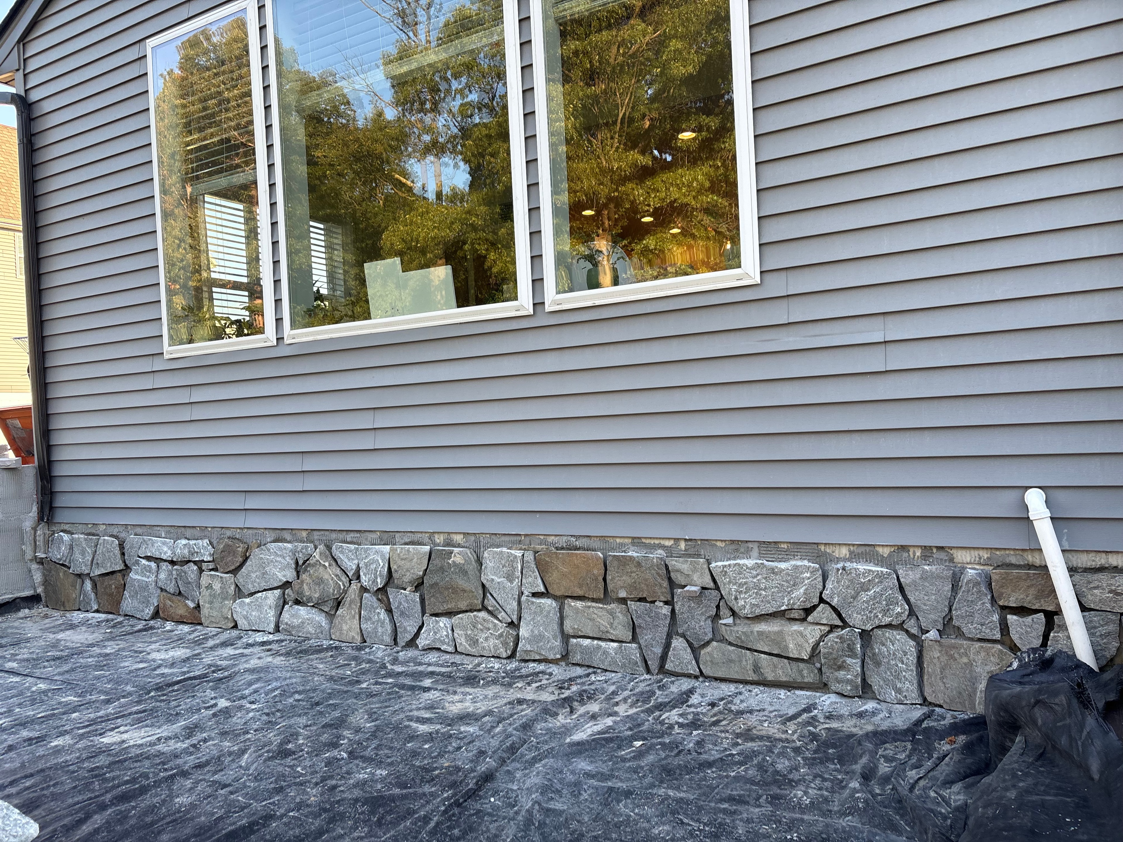 Chef's Dream — close-up of natural stone veneer being applied to base of house beside patio, landscape fabric and drain pipe visible, gray siding and large windows above
