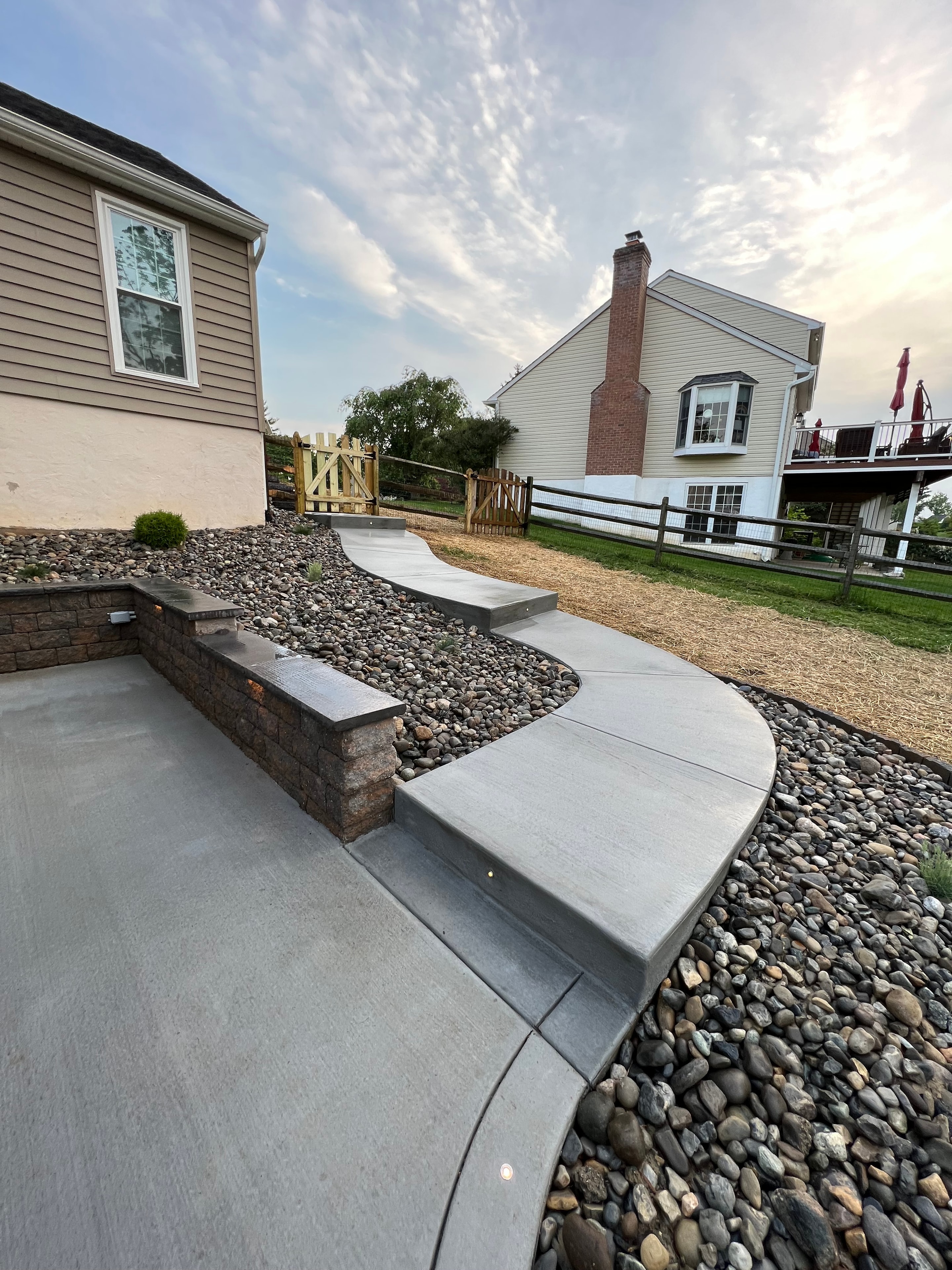 Concrete Jungle — daytime view of freeform concrete patio with curved retaining wall, river rock border, LED step lights, and wooden gate in background