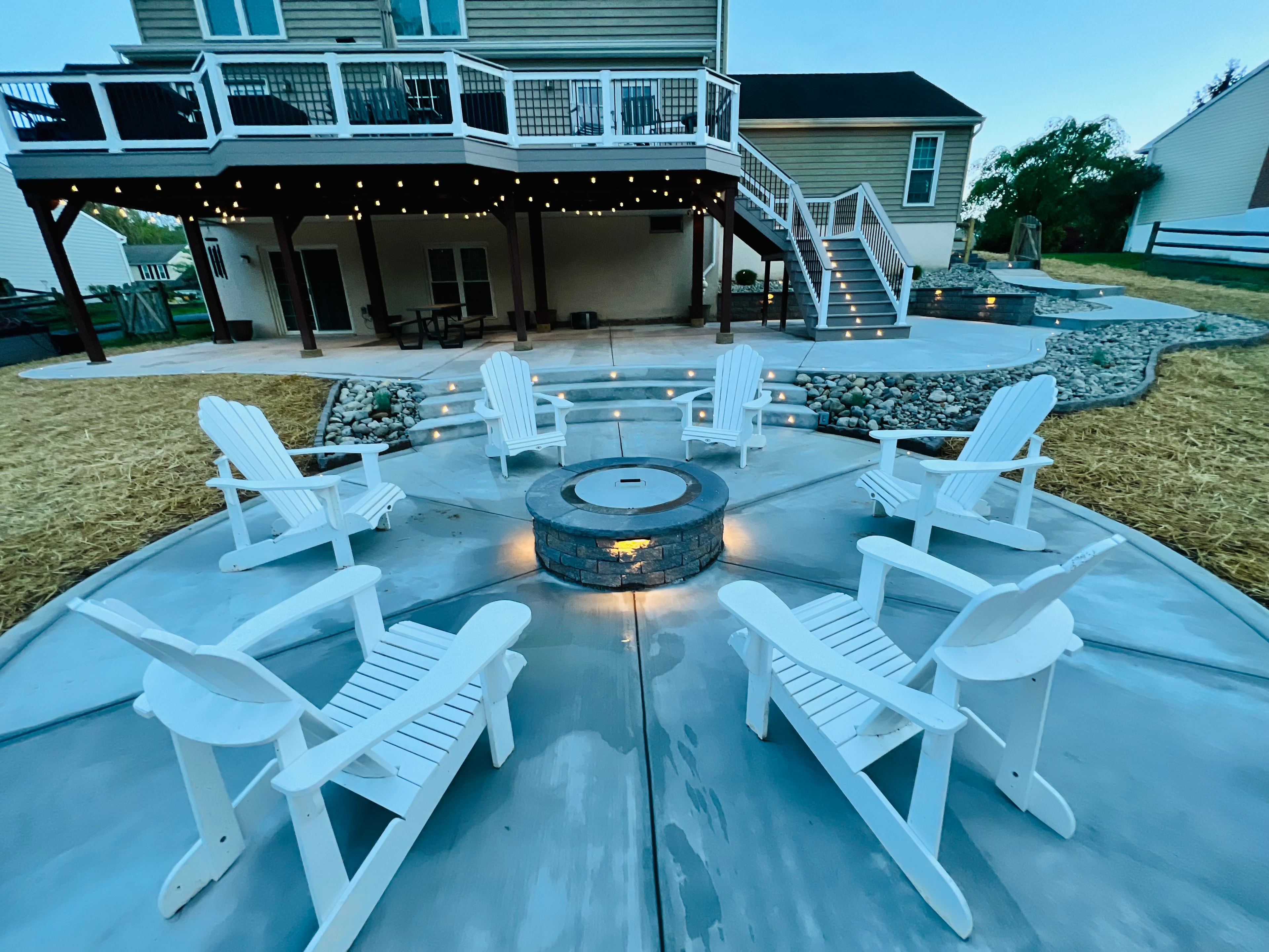 Concrete Jungle — dusk wide-angle shot of circular fire pit with white Adirondack chairs surrounding it, LED step lights, string lights, and full multi-level deck backdrop