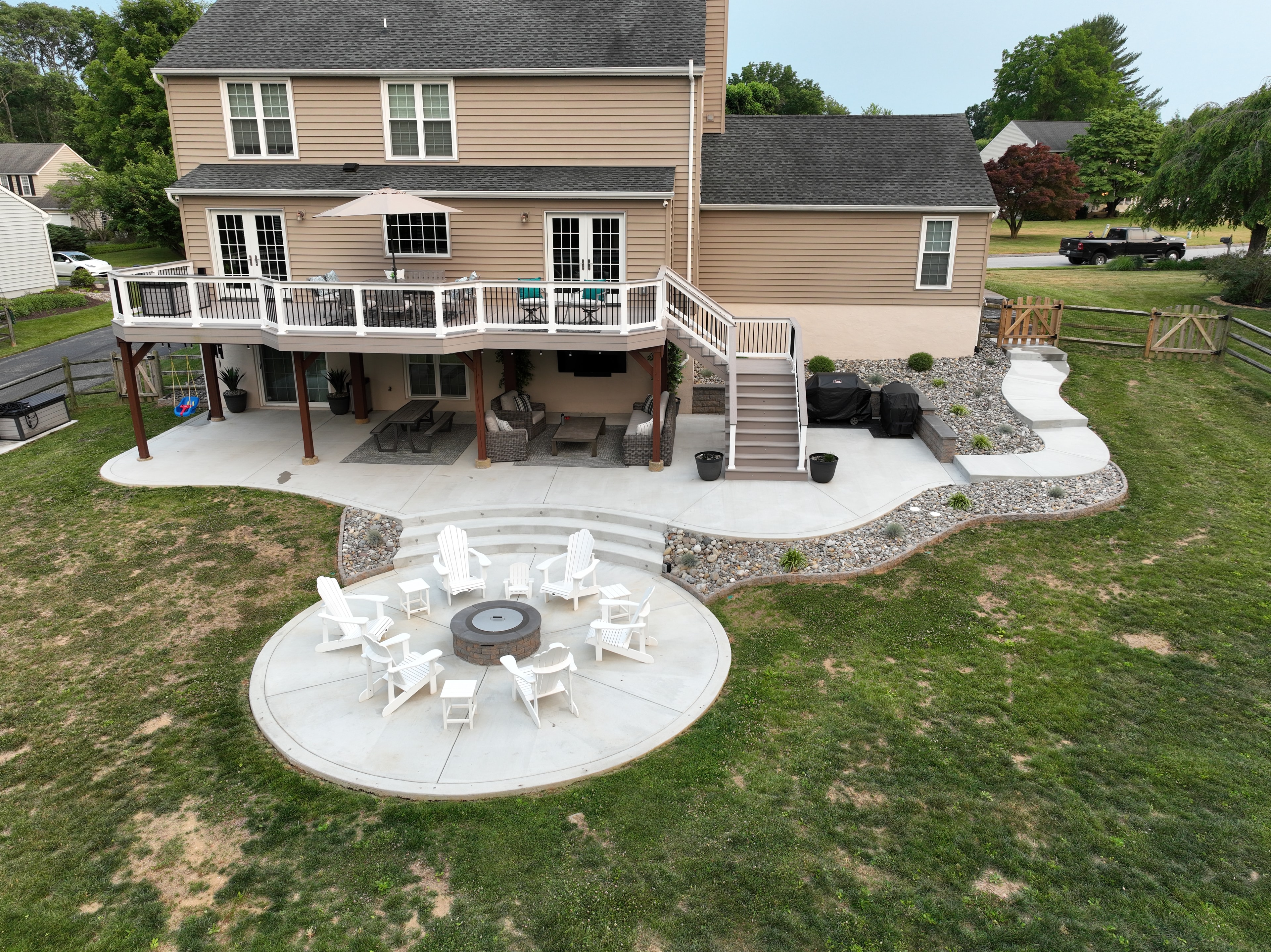 Concrete Jungle — aerial dusk angled shot of freeform concrete patio with circular fire pit seating area, multi-level deck with LED step lights, under-deck living space, and river rock border