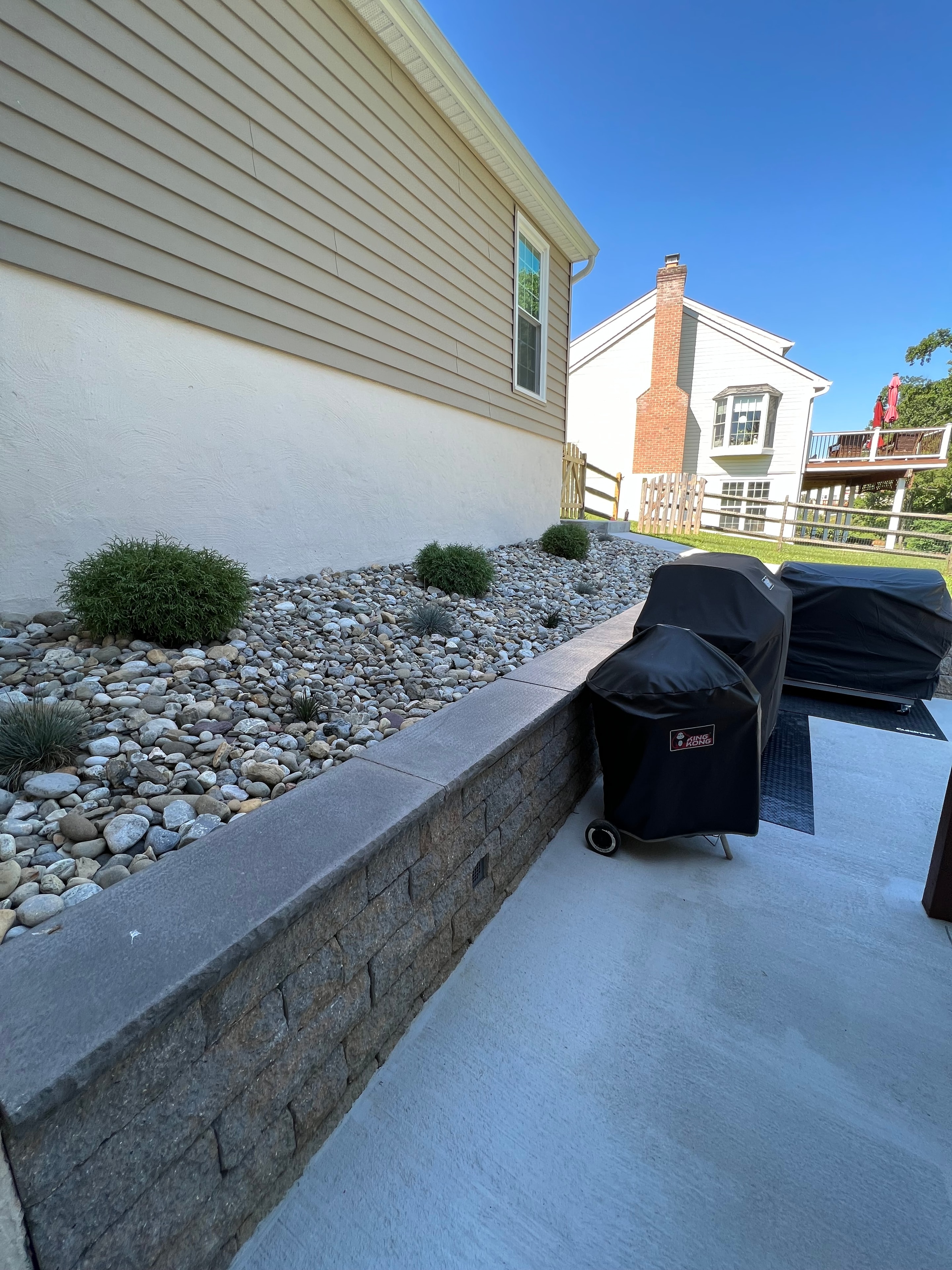 Concrete Jungle — daytime close-up of granite sitting wall with river rock landscaping, grill station, and ornamental shrubs beside house