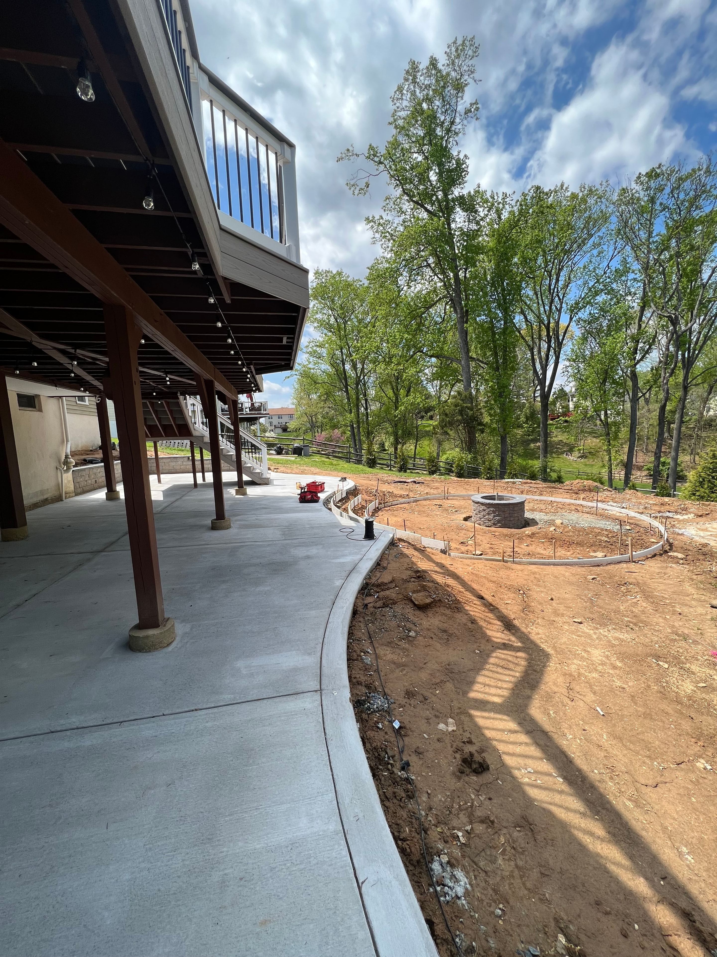 Under construction — view from multi-level deck staircase showing fresh concrete patio, circular fire pit form in bare dirt, deck railing and shed visible