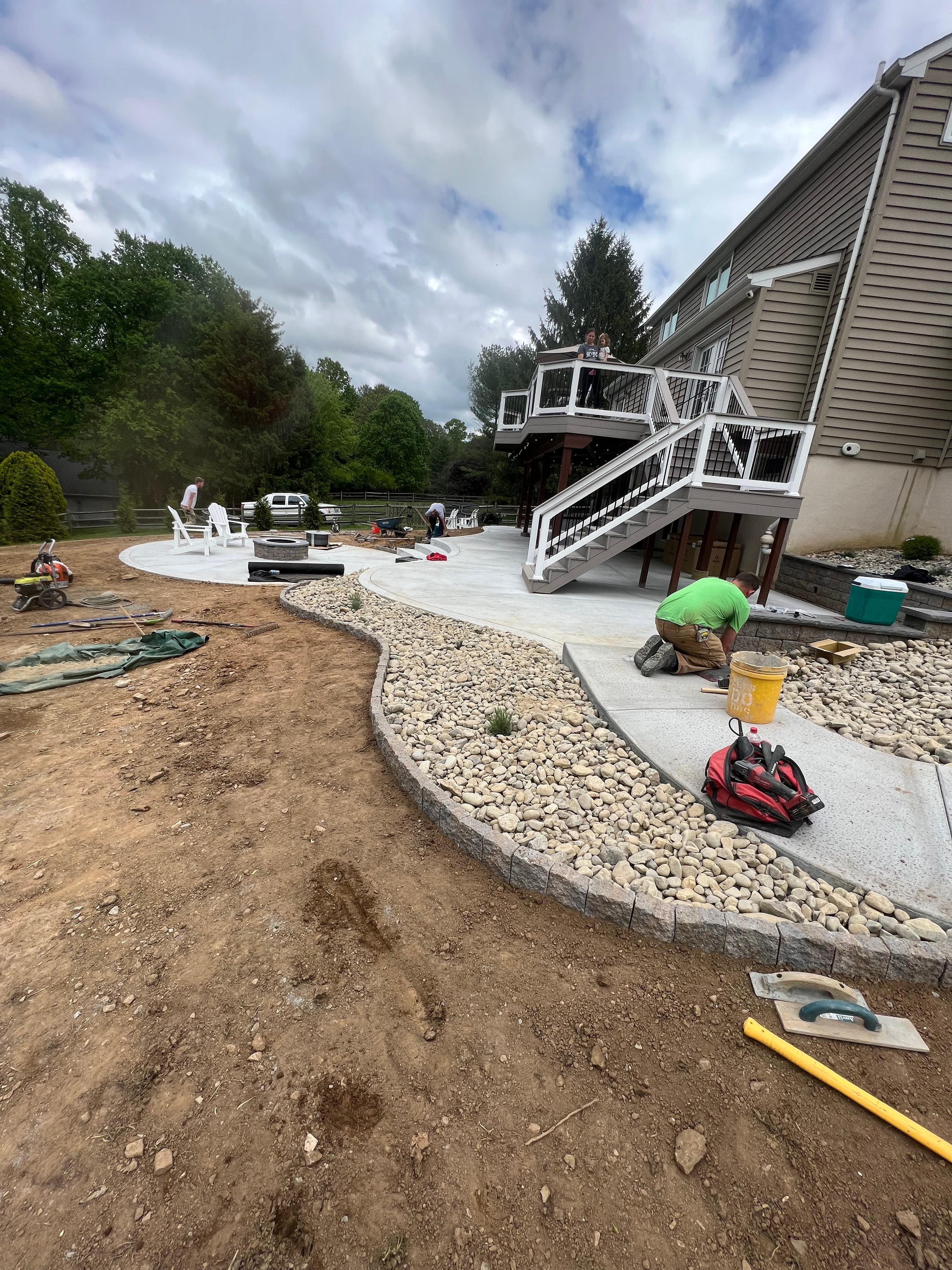 Under construction — crew member in green shirt setting river rock border along curved concrete patio edge, tools and yellow float on site, multi-level deck in background