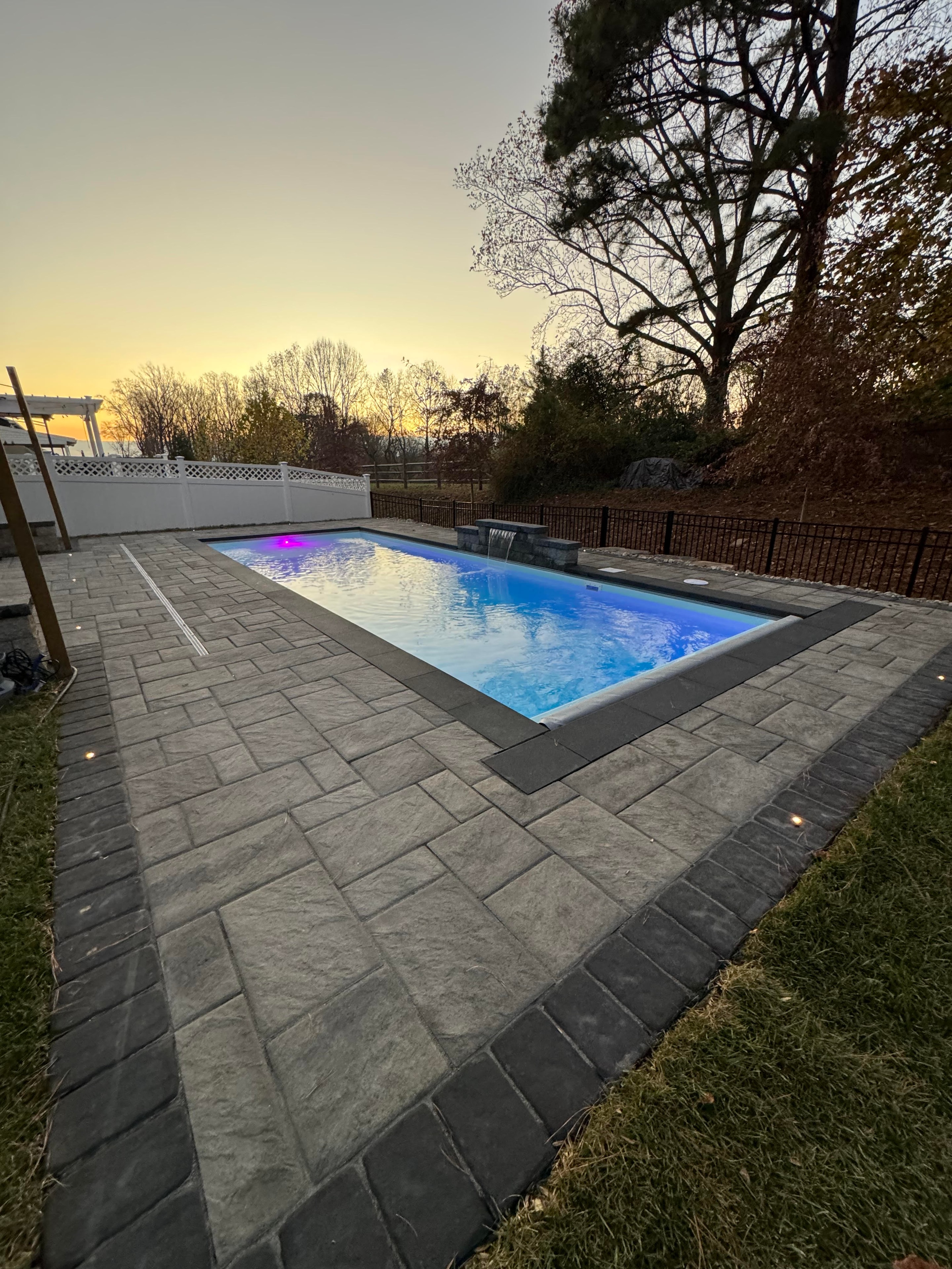 Small but Mighty — wide dusk shot of pool with paver patio, waterfall, and sunset treeline