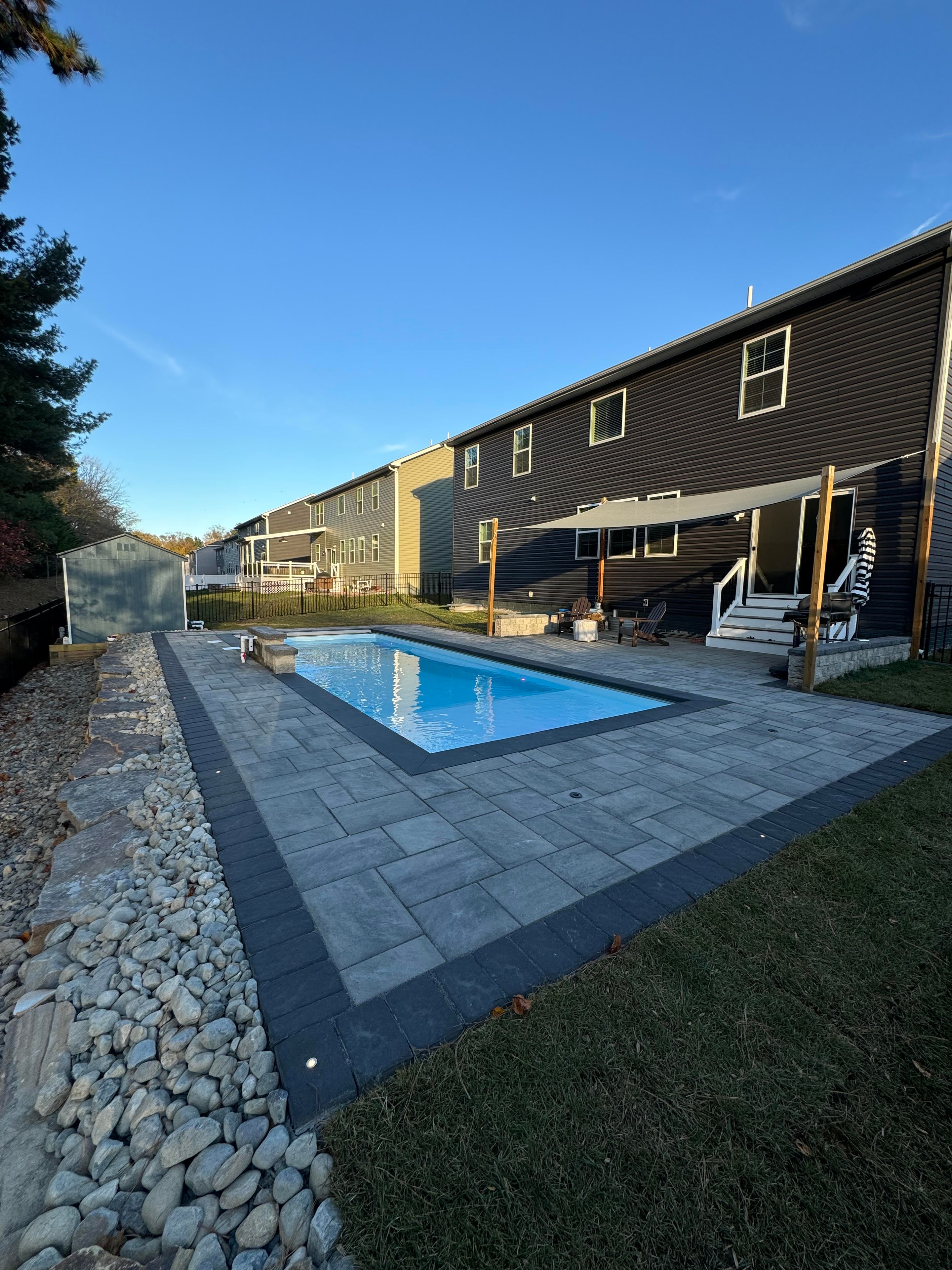 Small but Mighty — daytime wide shot of pool with paver surround, shade sail, and dark-sided house