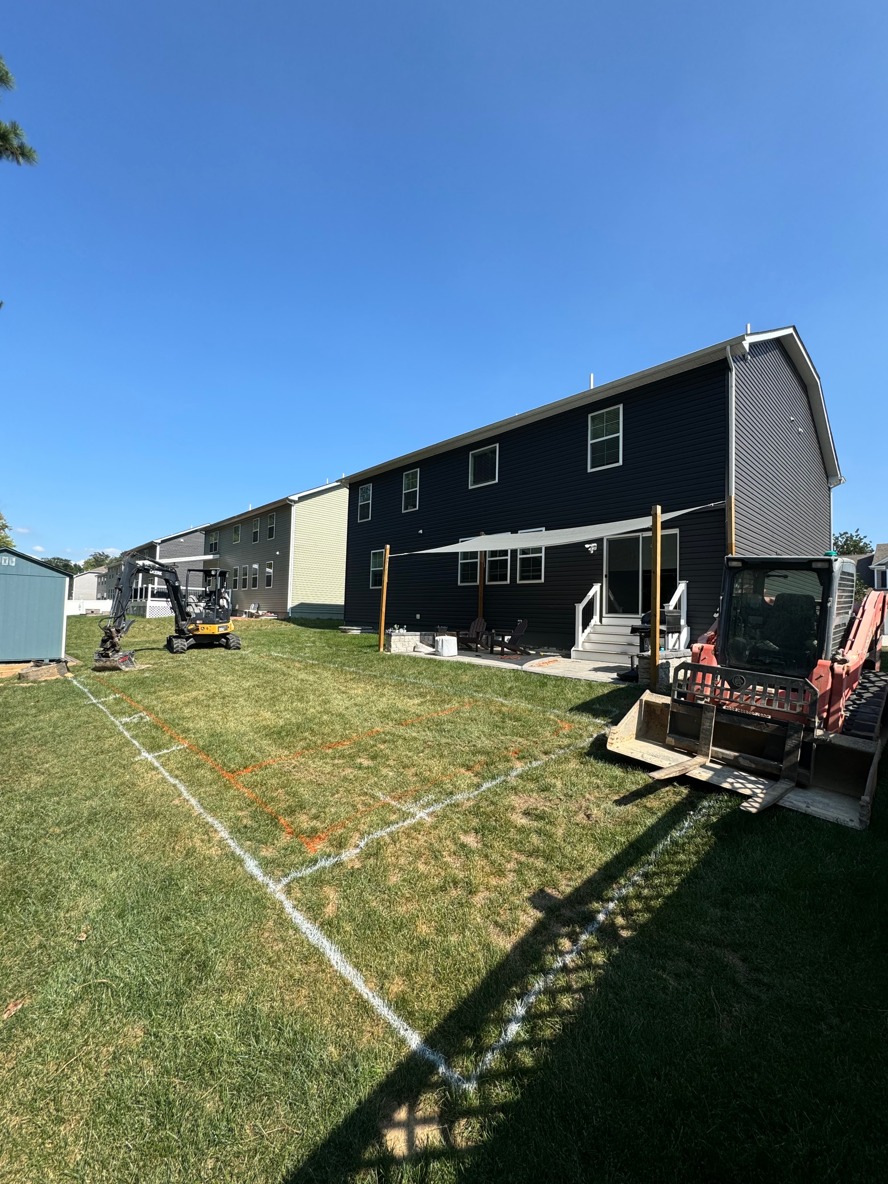 Under construction — backyard staked out with pool area marked, excavator and equipment on site beside dark-sided house