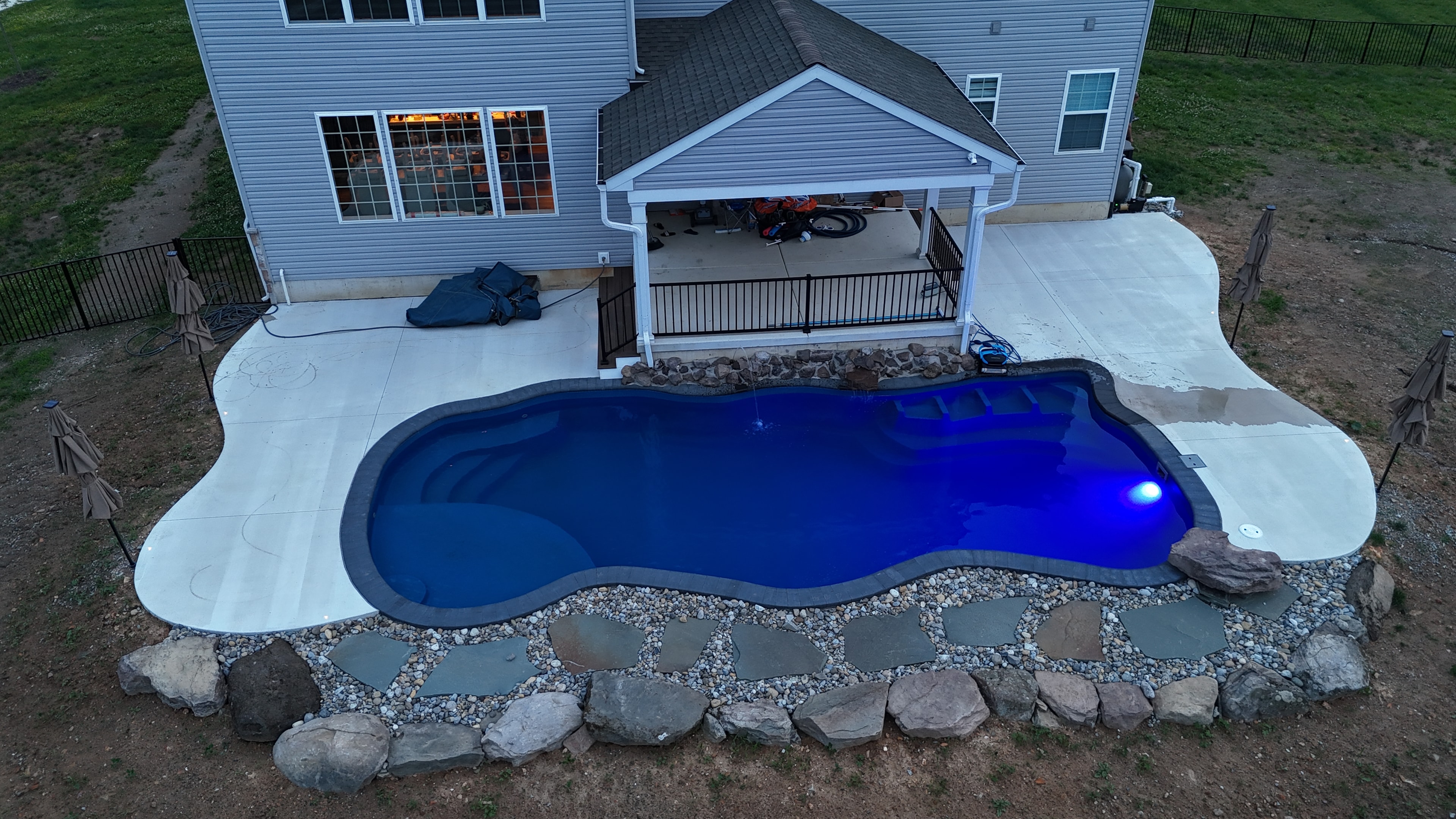 Tight Quarters — aerial daytime shot of pool with concrete surround, pergola, rock border, and neighboring homes
