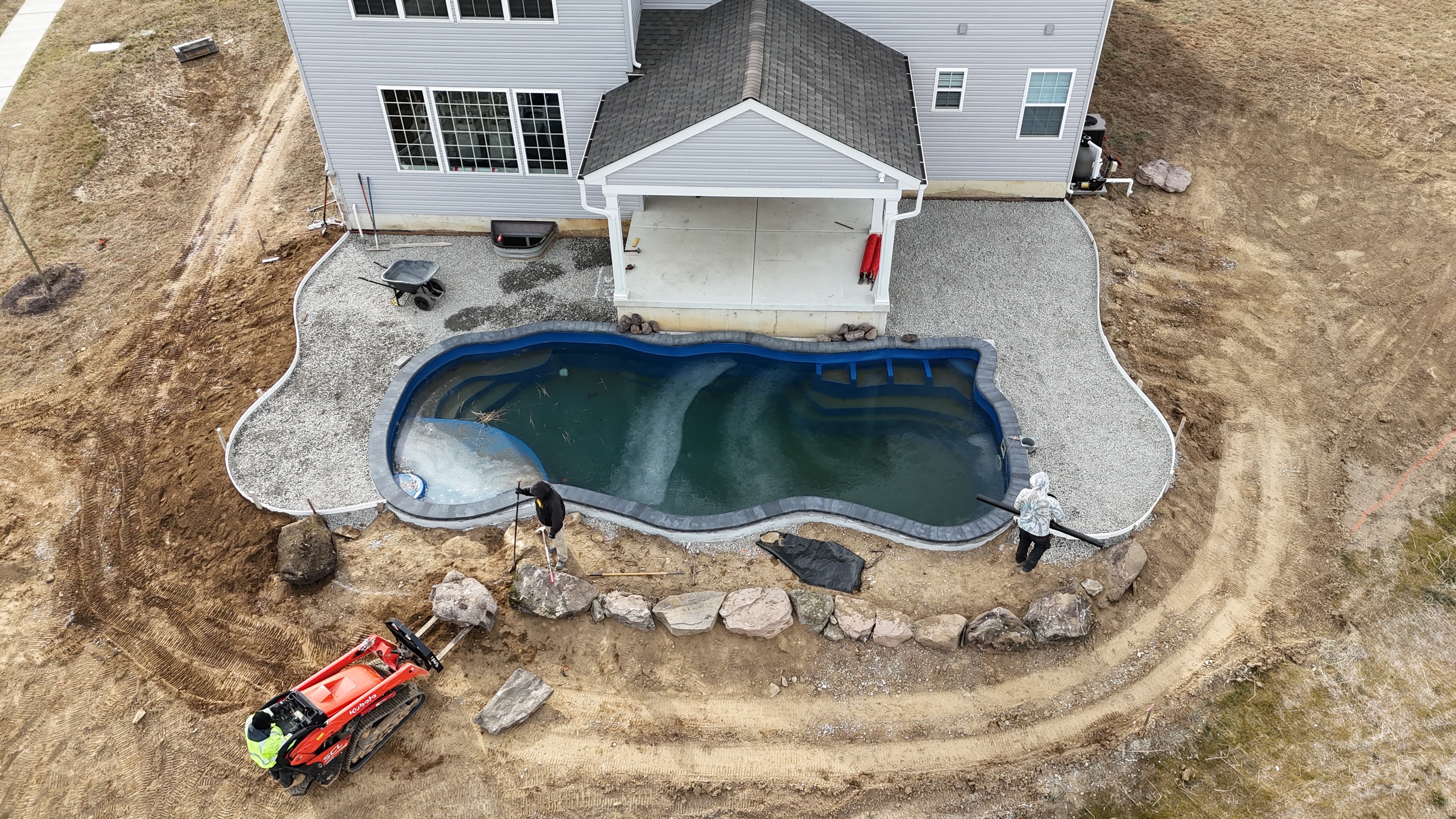 Under construction — aerial view of pool with coping installed, gravel base taking shape, crew and red Kubota skid steer placing boulders around perimeter