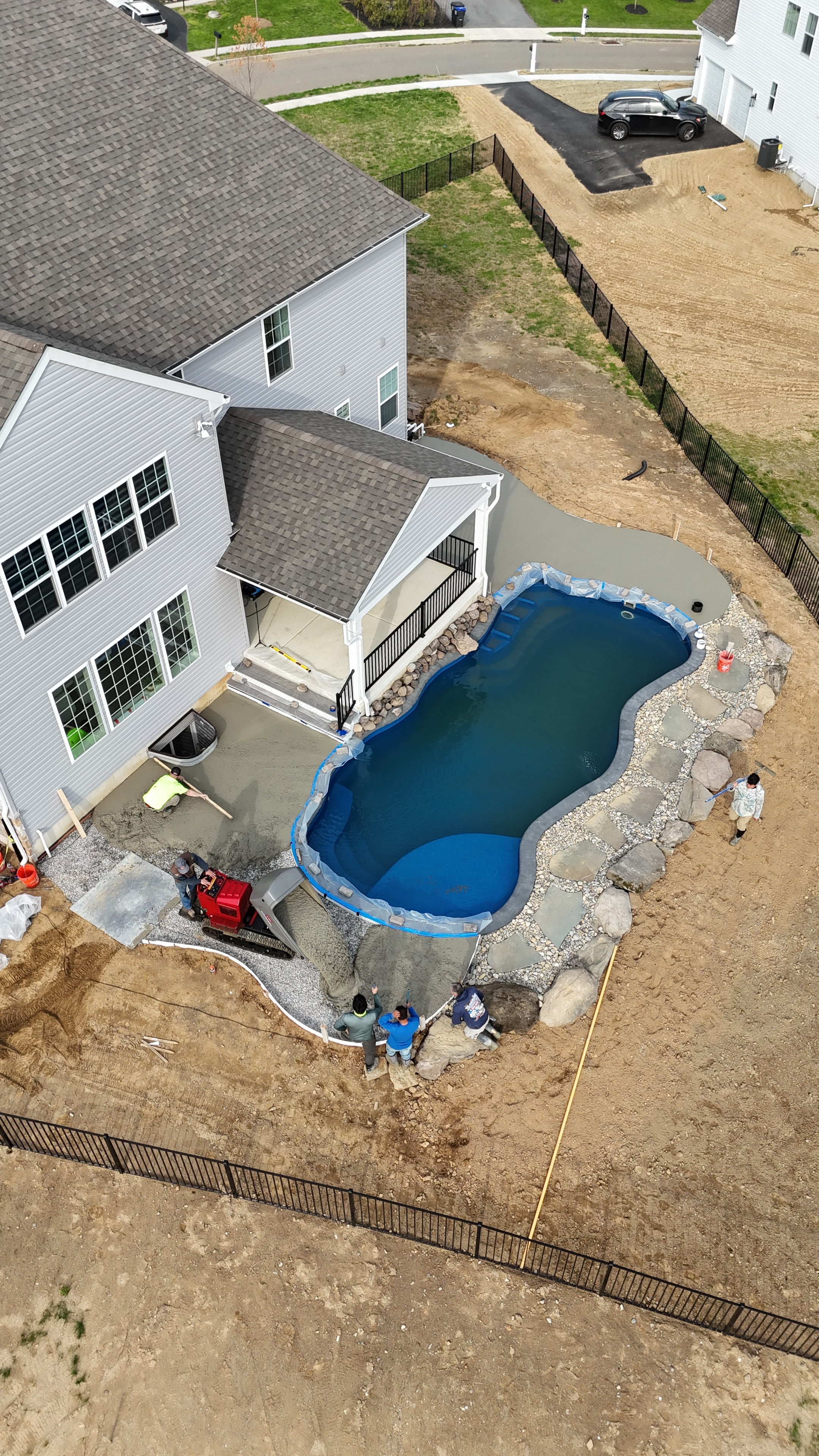 Under construction — aerial angled shot of pool with concrete patio and crew working, red Kubota skid steer on site, boulder and river rock border being set